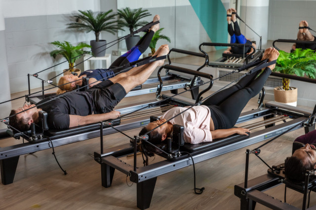Several individuals perform Pilates exercises, lying supine on reformers.