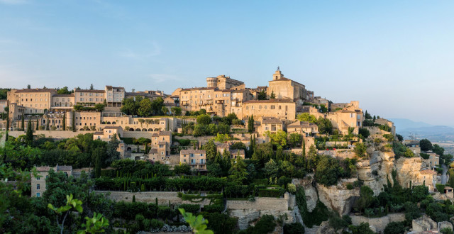 Stone buildings stand clustered atop a hill, surrounded by lush greenery and gardens, under a clear blue sky, overlooking a distant landscape of rolling hills