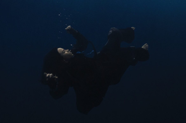 A person, dressed in dark clothing, is floating and slightly rotating underwater in a deep, shadowy environment, with bubbles rising around them.
