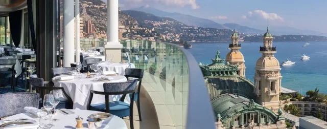 Restaurant tables are set with white tablecloths and glassware, positioned on a terrace overlooking coastal city architecture and the sea, with boats floating and mountains in the background.