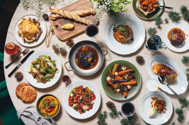 A variety of gourmet dishes are artfully arranged on a round table, featuring vibrant salads, roasted vegetables, and pastries, surrounded by decorative greenery and pine cones, creating a festive dining atmosphere.