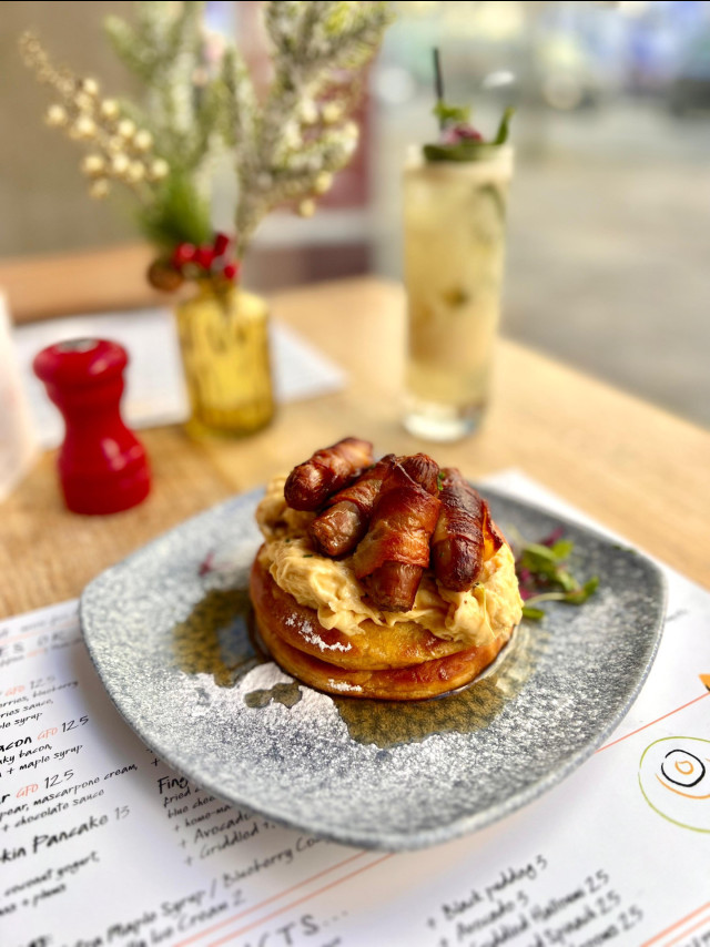 A stack of pancakes topped with bacon and sauce sits on a square plate. In the background, there's a drink and small vase on a wooden table with a blurred menu.