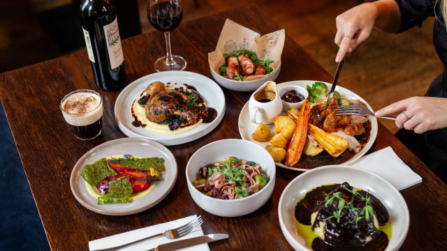 A variety of plated dishes on a wooden table, with a person using utensils. Surrounding are a wine bottle, glass, coffee, and napkin set, creating a dining atmosphere.