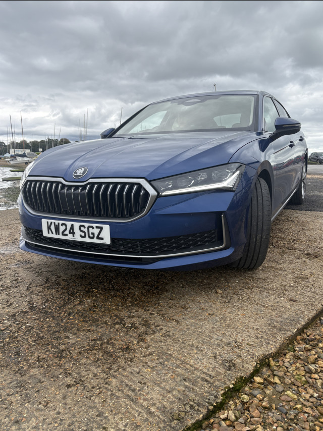 A blue car is parked on an uneven, textured surface near water and sailboats under a cloudy sky. The license plate reads “KW24 SGZ.”