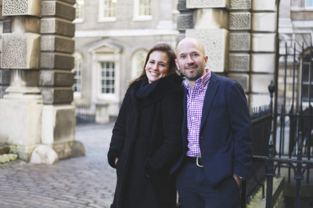 Two people stand smiling in a cobblestone courtyard, leaning casually against a black wrought iron railing, surrounded by historic stone buildings with large windows