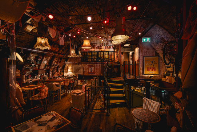 A cozy, dimly-lit pub features eclectic decor with vintage lamps, brick walls, and a spiral staircase. Various seating areas include checkered-cloth tables. A sign reads 