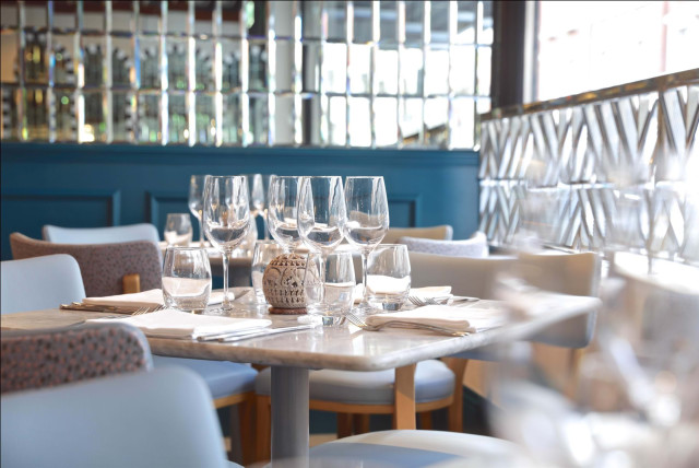 Wine glasses and water glasses are neatly arranged with napkins on a set dining table in a stylish restaurant, featuring upholstered chairs and decorative mirrors on blue walls