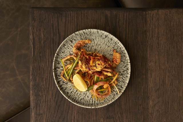 A plate holds a cooked crab garnished with green beans, red onions, and a lemon wedge, resting on a dark wooden table.