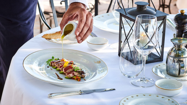 A hand is pouring sauce over a gourmet dish on an elegantly set table with a lantern, wine glass, and cutlery, creating an upscale dining atmosphere outdoors