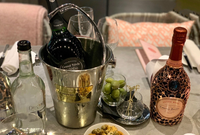A bottle of Grand Siècle champagne chills in an ice bucket on a dining table, accompanied by a pink Laurent-Perrier bottle, green olives, condiments, and folded napkins in a cozy setting