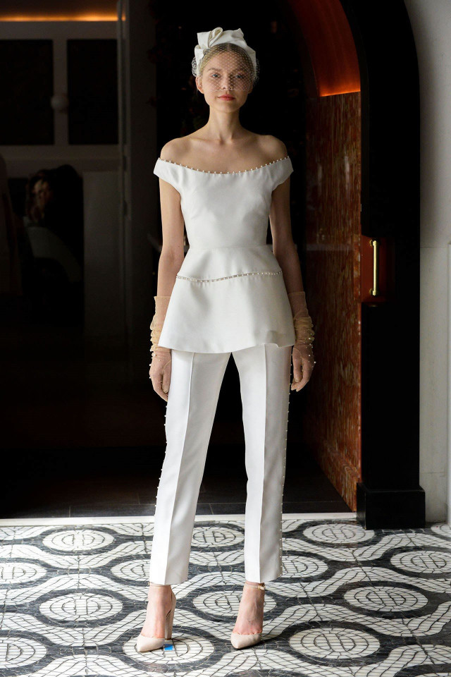 A woman stands wearing an elegant, off-shoulder white ensemble with a peplum top and pants, a headband with netting, and nude heels, on a patterned floor in an ornate hallway