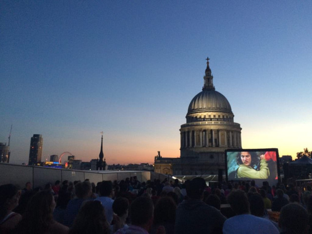 A crowd watches an outdoor movie on a large screen at dusk, with a historic domed building and city skyline in the background under a clear evening sky