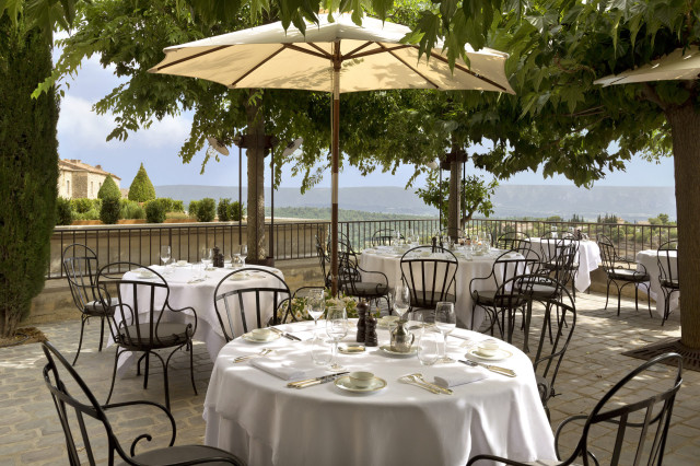 Tables with white tablecloths are set with dishes and glasses, arranged under large umbrellas on a stone patio, surrounded by trees and overlooking a scenic valley landscape