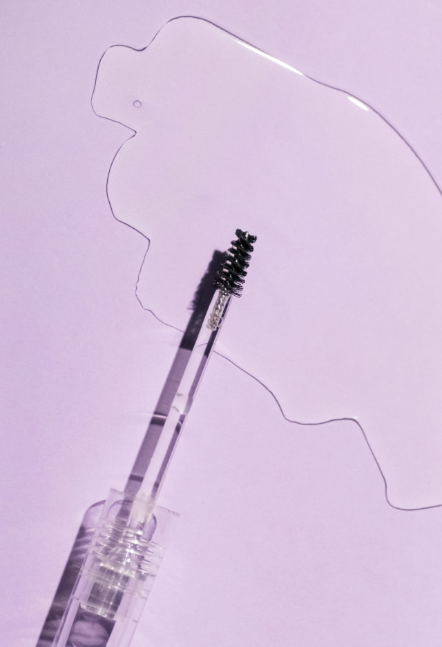 A clear mascara wand lies diagonally on a pastel purple surface, next to an amorphous, shiny liquid spill, casting soft shadows