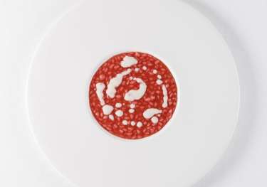 A white plate holds a circular slice of red salami with white fat specks, set against a plain white background.