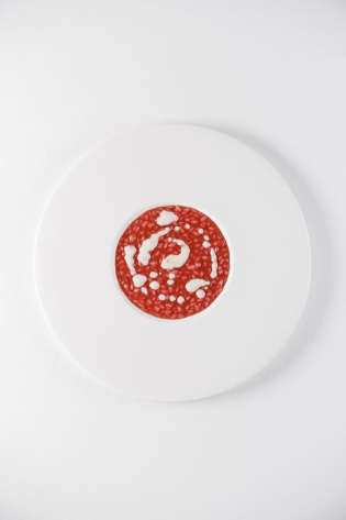 A white plate holds a circular slice of red salami with white fat specks, set against a plain white background.