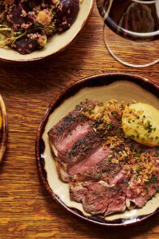 A wooden table holds assorted dishes: sliced steak with garnish, hearty stew, fresh vegetable salad, quinoa mix, and a glass of red wine, creating a rich dining scene.