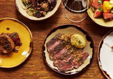 A wooden table holds assorted dishes: sliced steak with garnish, hearty stew, fresh vegetable salad, quinoa mix, and a glass of red wine, creating a rich dining scene.