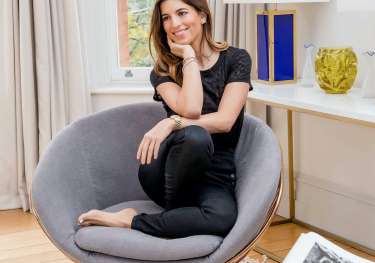 A woman sits cross-legged on a modern gray chair, smiling thoughtfully.