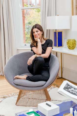 A woman sits cross-legged on a modern gray chair, smiling thoughtfully.