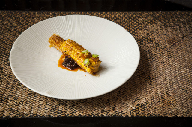 Grilled corn coated with spices rests on a white plate, garnished with chopped green onions; the plate is set on a woven, neutral-toned placemat
