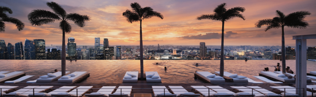 Palm trees line an infinity pool where people swim and relax on lounge chairs, overlooking a cityscape during a vibrant sunset sky