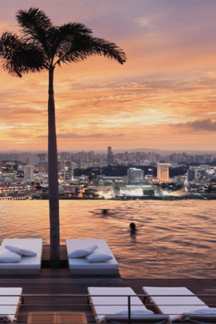 Palm trees line an infinity pool where people swim and relax on lounge chairs, overlooking a cityscape during a vibrant sunset sky.