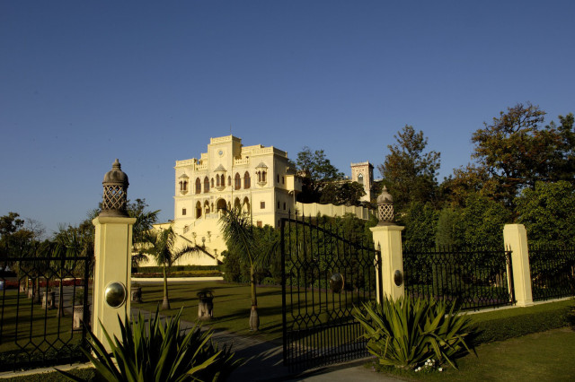 A large, elegant castle stands amidst lush gardens, bordered by decorative wrought-iron gates and flanked by trees, under a clear blue sky