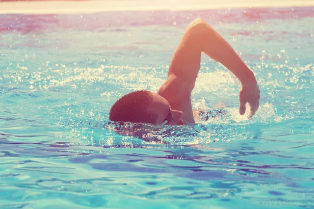 A person swims freestyle in a sunlit outdoor pool, creating splashes around them as they move through the bright blue water