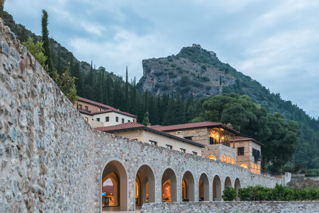 Stone building with arched walkways sits against a mountain backdrop, surrounded by lush greenery and trees, under a cloudy sky