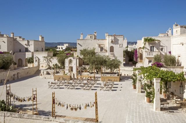 A rustic courtyard features wooden tables and chairs arranged under olive trees, surrounded by white stone buildings adorned with vines and flowers, under a clear blue sky