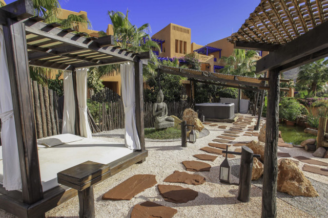 A wooden canopy bed with white curtains stands under a pergola, surrounded by stone pathways and lush greenery, with a Buddha statue and hot tub in the background