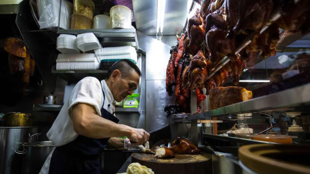 A chef slices roasted duck, surrounded by hanging meats and stacked dishes in a small, well-lit food stall kitchen