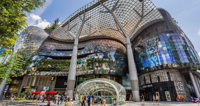 Shoppers stroll in front of a modern, glass-facade shopping mall featuring luxury brand stores, such as Louis Vuitton and Giorgio Armani, under a unique, curvilinear canopy structure on a sunny day