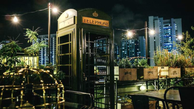 A vintage green telephone booth stands unused on a rooftop garden at night, surrounded by plants and wooden crates, with city skyscrapers illuminated in the background