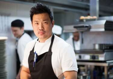 Chef stands confidently, wearing a white shirt and black apron, in a busy kitchen with chefs behind him and cooking equipment around.
