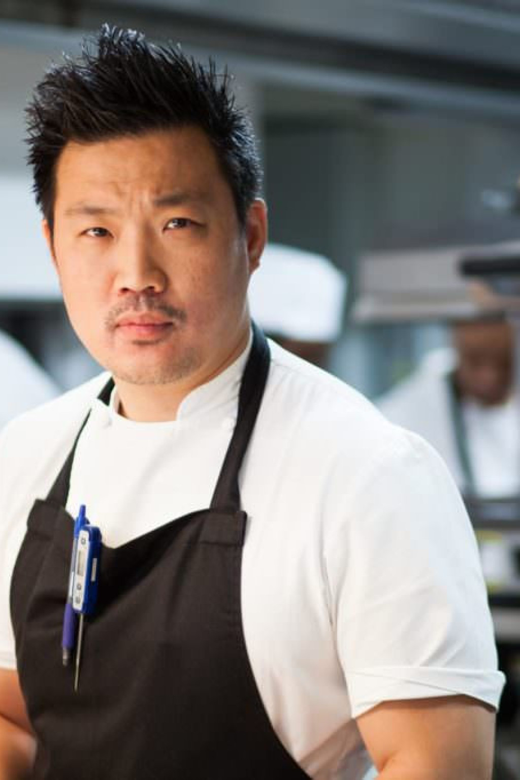 Chef stands confidently, wearing a white shirt and black apron, in a busy kitchen with chefs behind him and cooking equipment around