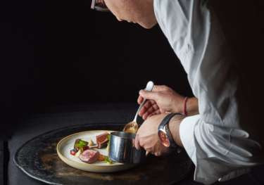 A chef meticulously garnishes a plated dish with sauce from a small pot, under soft lighting, focused on precision and presentation in an elegant, dark culinary setting.