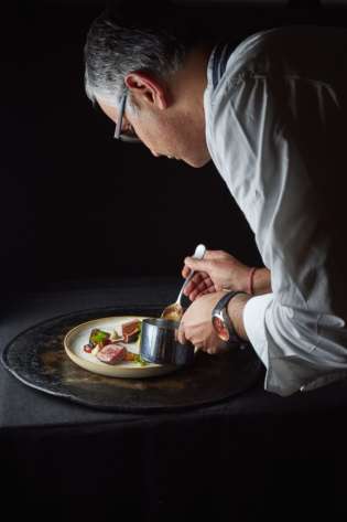 A chef meticulously garnishes a plated dish with sauce from a small pot, under soft lighting, focused on precision and presentation in an elegant, dark culinary setting.