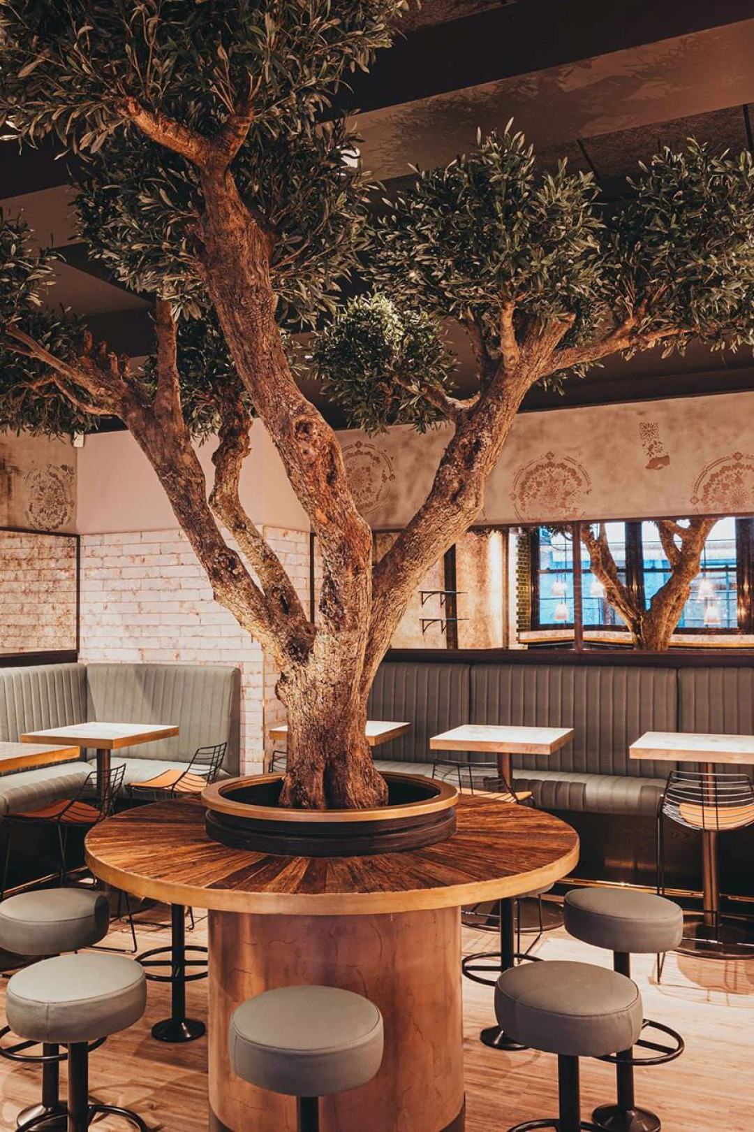 A large tree grows through the center of a round table, surrounded by stools in a cozy restaurant setting with brick walls and windows