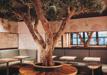 A large tree grows through the center of a round table, surrounded by stools in a cozy restaurant setting with brick walls and windows.