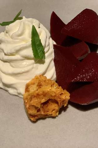 A dessert plate features whipped cream with green leaf garnish, alongside sliced red fruit and a piece of honeycomb, set on a neutral-colored plate.