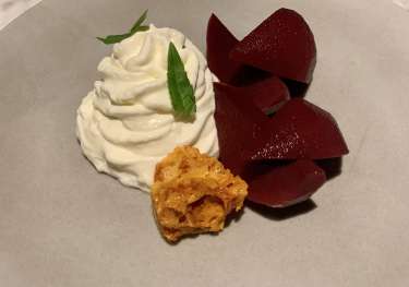 A dessert plate features whipped cream with green leaf garnish, alongside sliced red fruit and a piece of honeycomb, set on a neutral-colored plate.