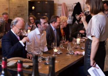 People seated around a wooden table engage in a wine tasting.