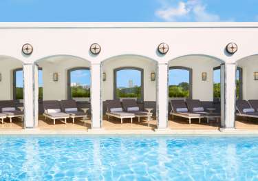 Poolside lounge chairs rest under white arched cabanas, with a blue pool in the foreground and greenery in the background, under a clear blue sky.