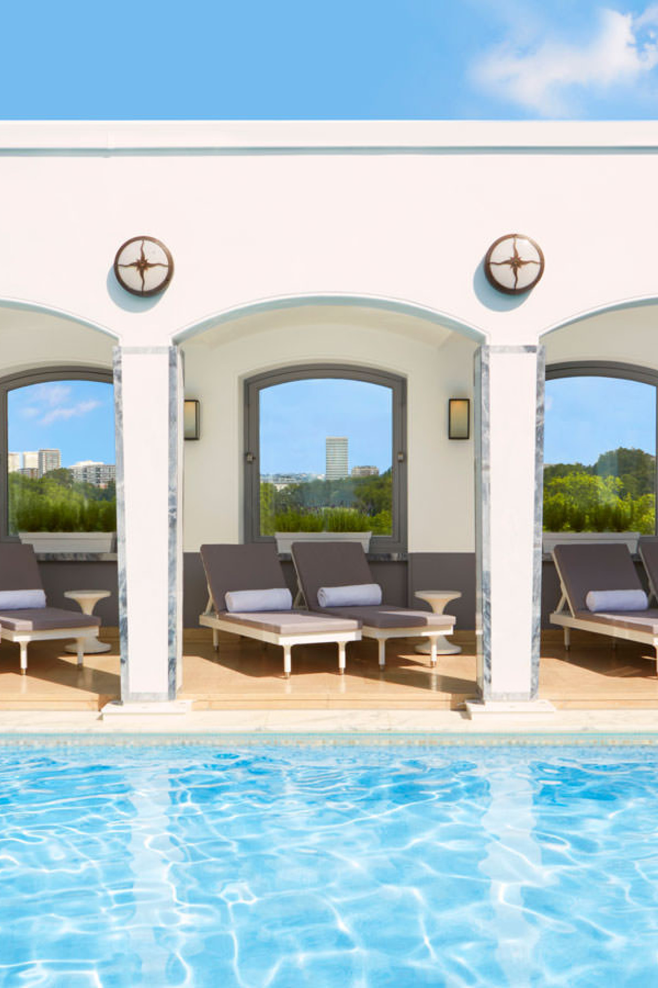 Poolside lounge chairs rest under white arched cabanas, with a blue pool in the foreground and greenery in the background, under a clear blue sky