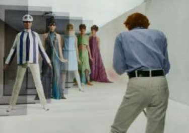 Man photographing mannequins in 1960s fashion in a studio.
