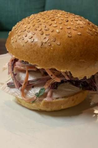 A sesame seed bun holds a sandwich filled with sliced meat, shredded vegetables, and dressing, resting on a white plate.
