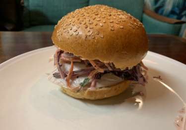 A sesame seed bun holds a sandwich filled with sliced meat, shredded vegetables, and dressing, resting on a white plate.