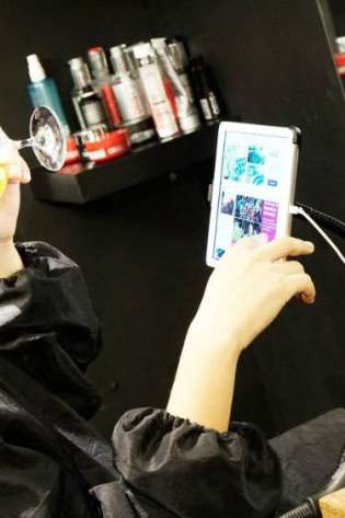A person in a barber's chair drinks beer while viewing a tablet.